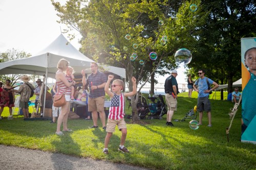 A child in sunglasses dances joyfully among bubbles at an outdoor event, with families and tents in the background.