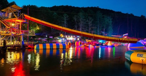 Colorful inflatable water attractions and a zip line illuminated at night, surrounded by trees and a calm lake.