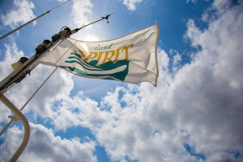 A flag with "Portland Spirit" waves against a backdrop of blue sky and fluffy clouds.