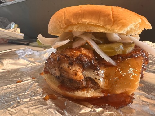 A close-up of a juicy burger with grilled chicken, pickles, onions, and barbecue sauce on a soft bun, wrapped in foil.