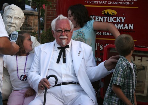 A man in a white suit and glasses sits with children, posing near a statue and a red truck at an outdoor event.