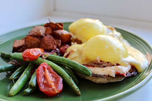 A green plate with eggs Benedict, roasted potatoes, green beans, and cherry tomatoes.