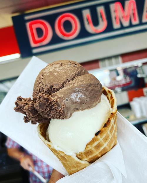 A hand holding a waffle cone with two scoops of ice cream: chocolate on top and vanilla below.