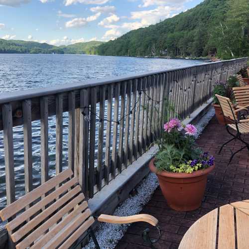 A serene lakeside view with wooden seating, potted plants, and mountains in the background under a blue sky.