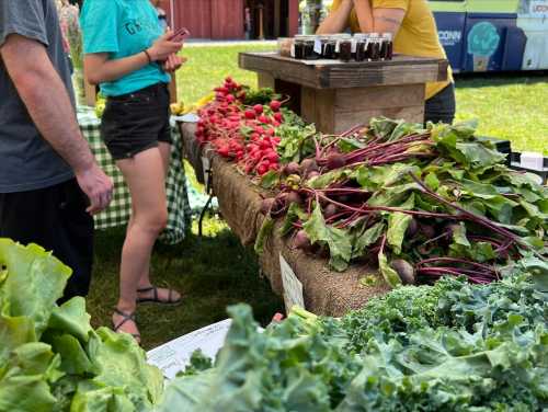 A vibrant farmers market display with fresh radishes, beets, lettuce, and kale, with people browsing nearby.