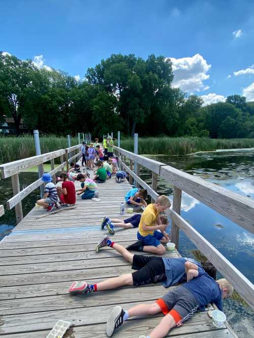 Children lying on a wooden dock, observing water and nature, with trees and a blue sky in the background.