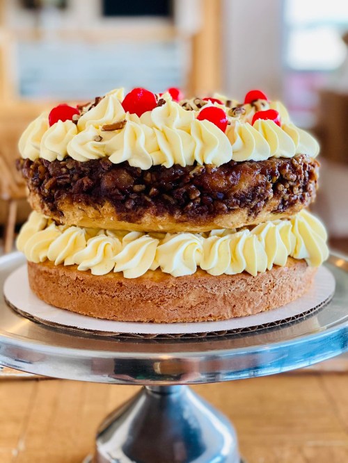 A layered cake topped with whipped cream, cherries, and a nutty caramel topping on a decorative stand.