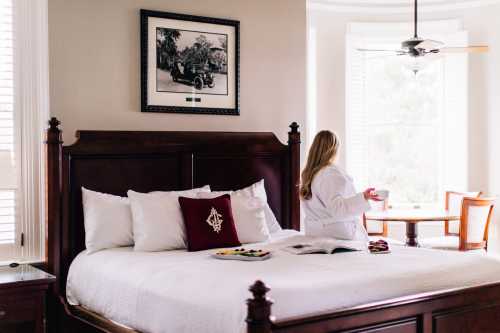 A woman in a robe sits on a bed, holding a cup, with a table and window in the background.