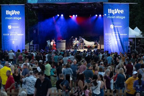 A large crowd enjoys a live band performance on the Hy-Vee main stage at an outdoor festival. Colorful lights illuminate the scene.