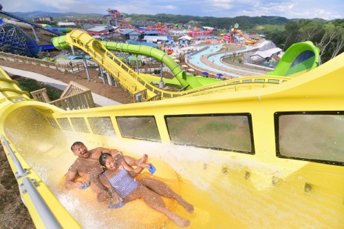 Two people slide down a yellow water slide, splashing into a pool at a water park with various attractions in the background.