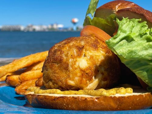 A close-up of a crab cake sandwich with lettuce and tomato, served with fries, against a scenic waterfront background.