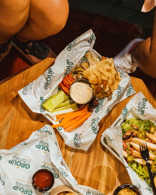 A wooden table with two trays of colorful food, including vegetables, pita, and dips, surrounded by people’s legs.