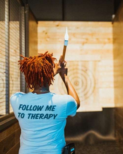 A person with orange dreadlocks stands in an axe-throwing venue, wearing a shirt that says "FOLLOW ME TO THERAPY."