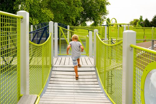 A child runs up a wooden ramp in a colorful playground surrounded by green trees and bright structures.