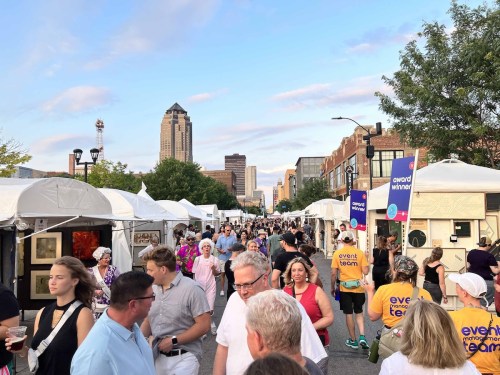 A bustling street fair with people browsing art booths, tents lined up, and city buildings in the background under a blue sky.