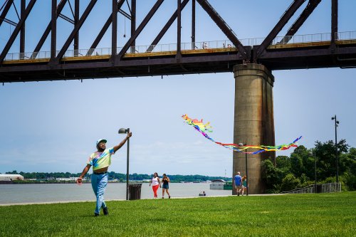 A person in a colorful outfit flies a kite under a bridge, with others enjoying the park by the water.