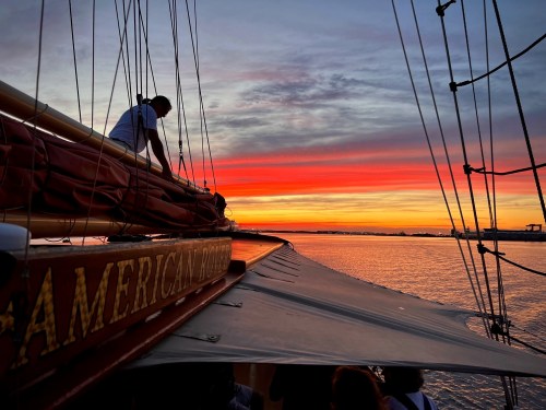 A sunset over water with vibrant orange and pink hues, viewed from a sailboat with a crew member adjusting the sails.