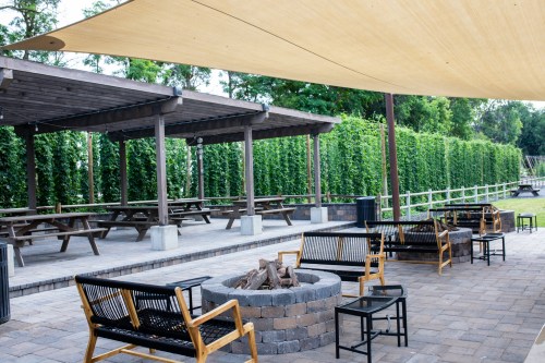 Outdoor seating area with a fire pit, shaded by a canopy, surrounded by greenery and picnic tables.