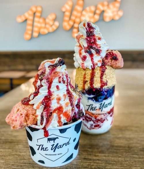 Two extravagant milkshakes topped with whipped cream, sauces, and cookies, displayed in front of a glowing sign.