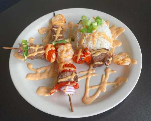 A plate of skewers with shrimp and beef, drizzled with sauce, served with a side of rice and green onions.