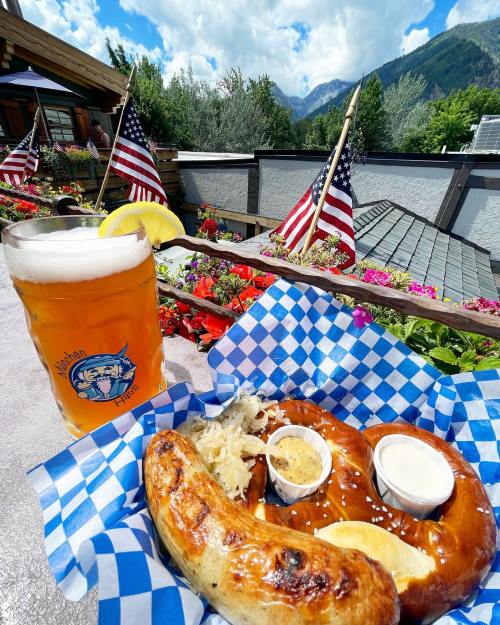 A pretzel and sausage with mustard and sauerkraut, alongside a beer, set against a scenic mountain backdrop.