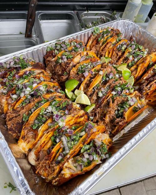 A tray of stacked tacos filled with meat, topped with onions, cilantro, and lime wedges.