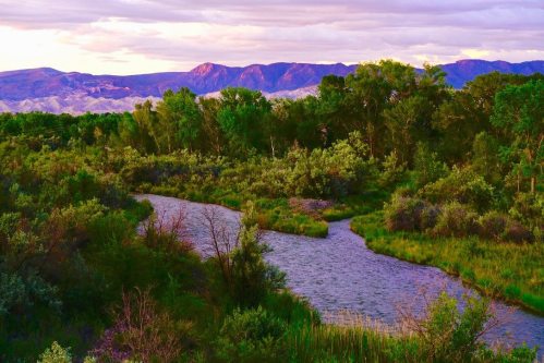 A winding river flows through lush greenery, with mountains in the background under a colorful sky at sunset.