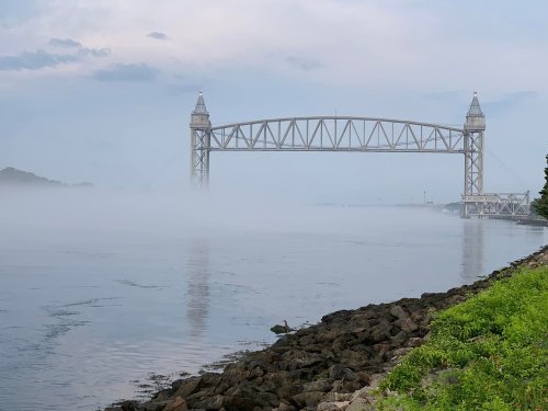 A foggy scene featuring a large metal bridge over a calm river, with rocky shores and greenery in the foreground.