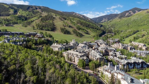 Aerial view of a mountain resort surrounded by lush green trees and rolling hills under a clear blue sky.