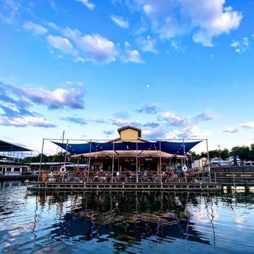 A waterfront restaurant with a blue canopy, surrounded by water and reflecting clouds in the sky.