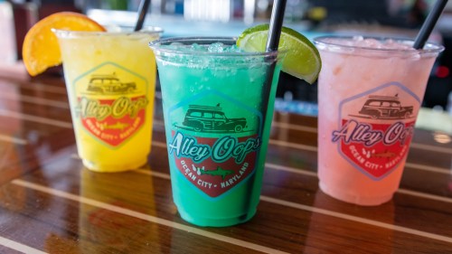 Three colorful drinks in clear cups with straws, featuring a yellow, green, and pink beverage, on a wooden bar.