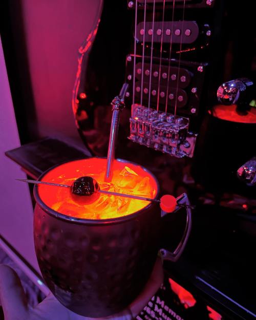 A hand holds a glowing cocktail with ice and a cherry, set against a backdrop of a guitar and vibrant lighting.