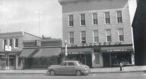 Black and white photo of a street scene featuring shops and a vintage car parked in front.