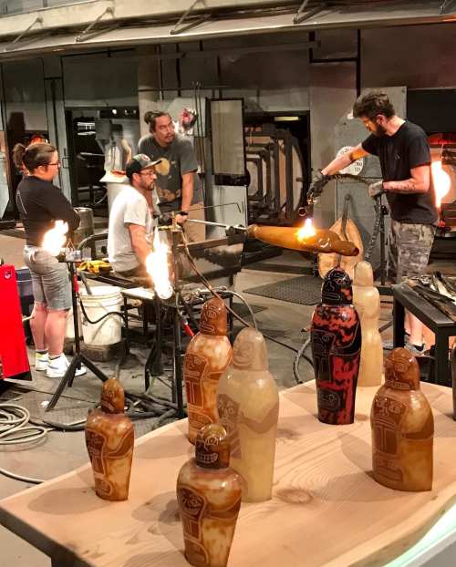 A glassblowing workshop with artists shaping molten glass, surrounded by finished glass sculptures on a table.
