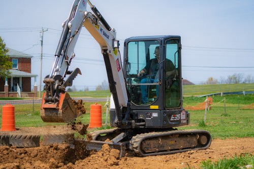 A small excavator digs into the ground on a construction site, with a house and green fields in the background.