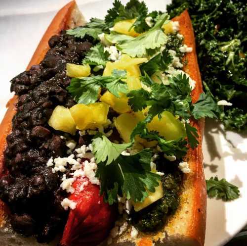 A gourmet hot dog topped with black beans, pineapple, cilantro, and cheese, served with a side of kale.