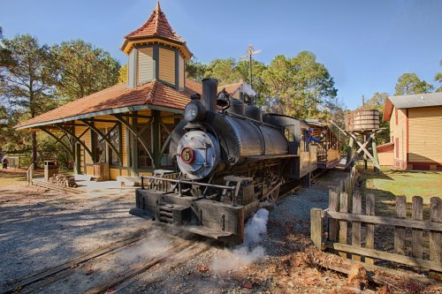 A vintage steam locomotive at a charming train station surrounded by trees and a wooden fence.