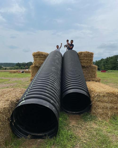 Two children play on large black pipes stacked on hay bales in a grassy field under a cloudy sky.