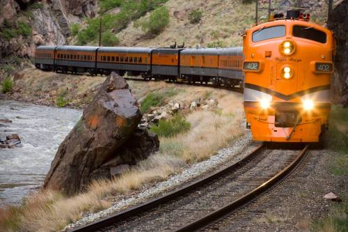 An orange train travels along a riverbank, surrounded by rocky terrain and greenery.