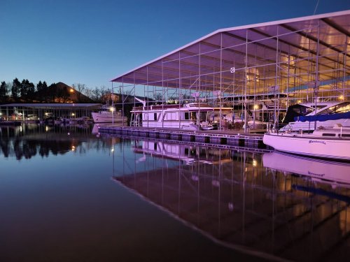 A serene marina at dusk, with boats docked and their reflections shimmering on calm water under a purple sky.