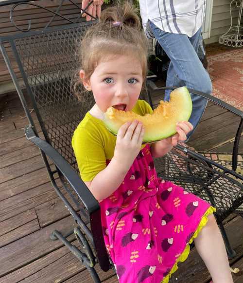 A young girl in a colorful dress sits on a chair, happily eating a large piece of melon.