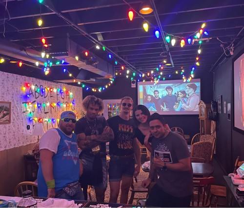 A group of five people poses in a colorful room decorated with string lights, with a retro movie playing in the background.