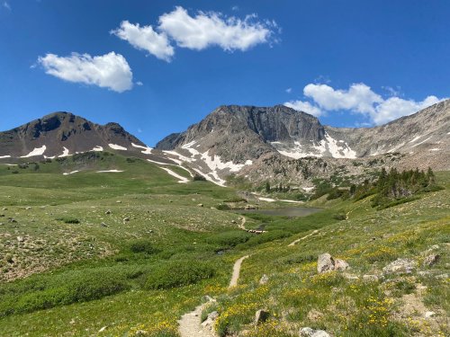 A scenic mountain landscape with green meadows, a winding path, and snow-capped peaks under a blue sky with clouds.