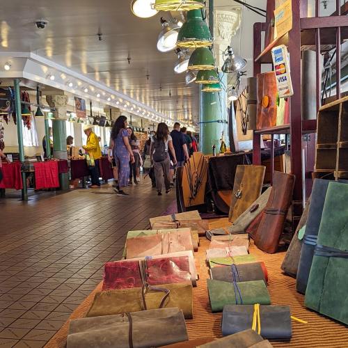 A vibrant market scene with colorful handmade journals displayed in the foreground and shoppers browsing in the background.