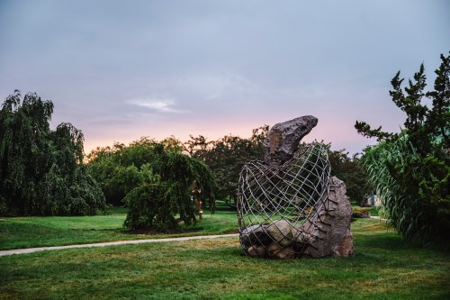 A large, abstract sculpture made of stone and wire sits in a grassy park, surrounded by trees under a cloudy sky.