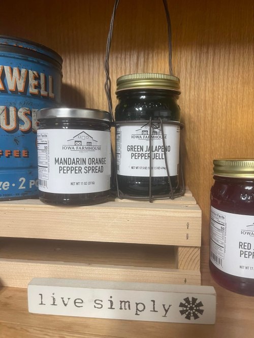 Two jars of pepper spreads on a wooden shelf, labeled Mandarin Orange and Green Jalapeño, with a sign that says "live simply."