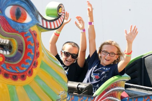 Two children with sunglasses and smiles raise their hands in excitement while riding a colorful amusement park attraction.