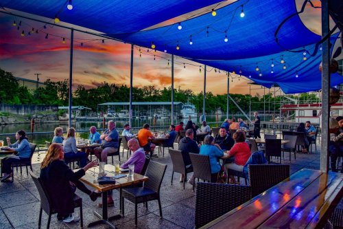 Outdoor dining area by the water at sunset, with people enjoying meals under a blue canopy and string lights.
