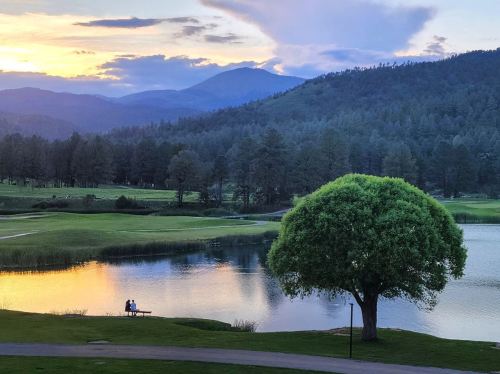 A serene landscape at sunset, featuring a lake, lush greenery, and two people sitting by the water's edge.