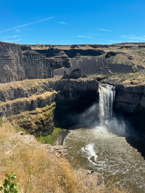 A waterfall cascades into a rocky canyon, surrounded by dry grass and steep cliffs under a clear blue sky.